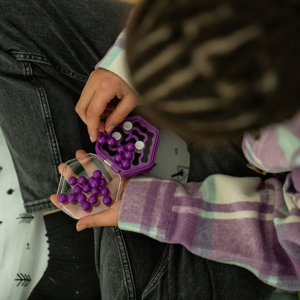 Child in a plaid shirt holding a purple Smart Games IQ Mini Hexpert puzzle on their lap, carefully placing spherical pieces into the transparent hexagon case.
