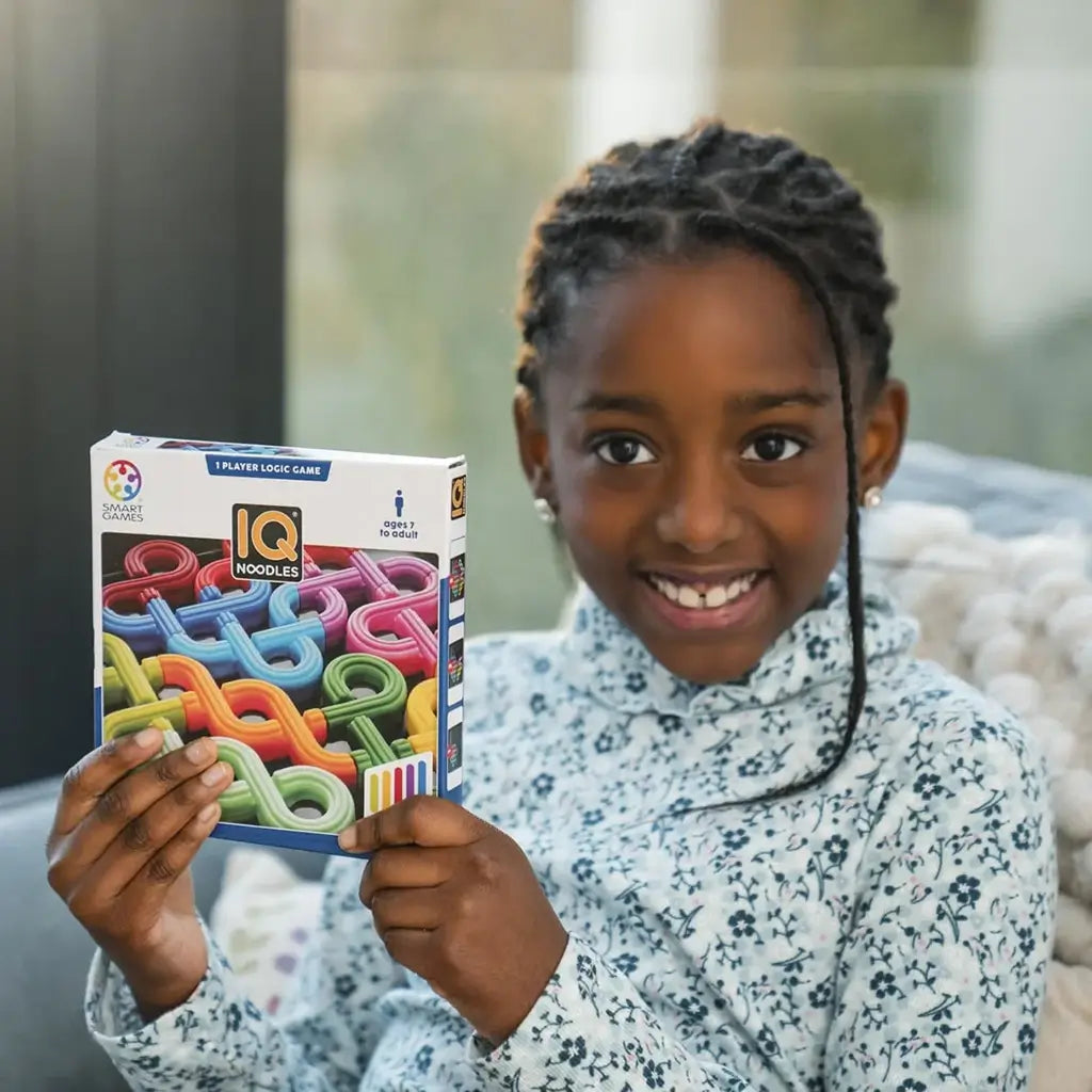 Smiling young girl in patterned shirt holds up the SmartGames IQ Noodles puzzle box, showing bright twisted noodle-shaped pieces in rainbow colors.