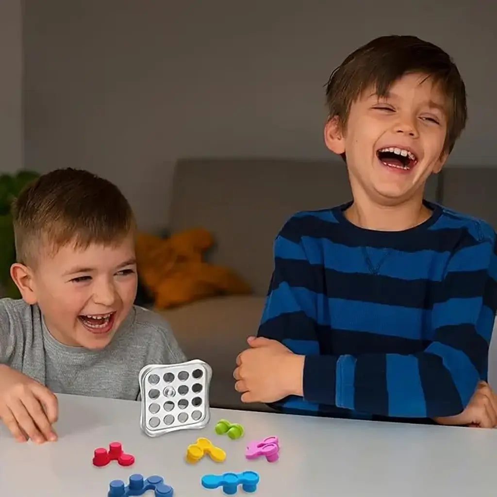 Two laughing boys sit at a table playing the Reverso Fidget Puzzle, with colorful game pieces spread out in front of the silver grid case during playtime.
