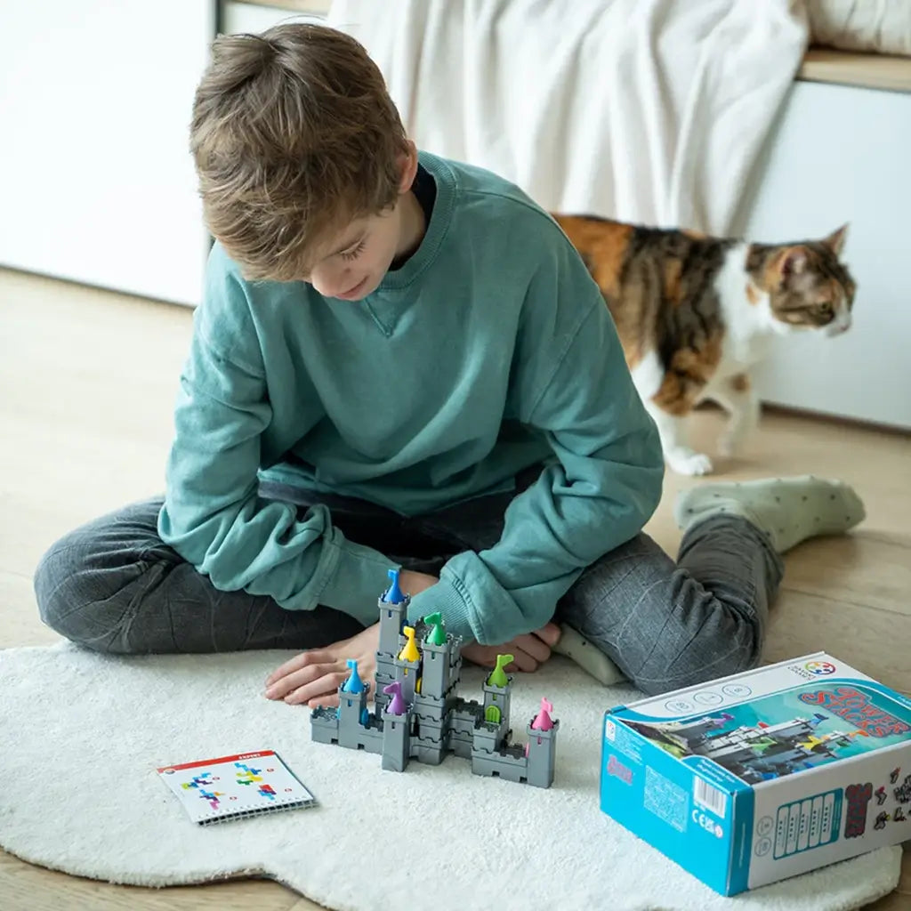 Boy in teal sweater plays Smart Games Tower Stacks on a white rug, assembling gray castle towers with colorful flags while a cat walks nearby.