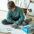 Boy in teal sweater plays Smart Games Tower Stacks on a white rug, assembling gray castle towers with colorful flags while a cat walks nearby.