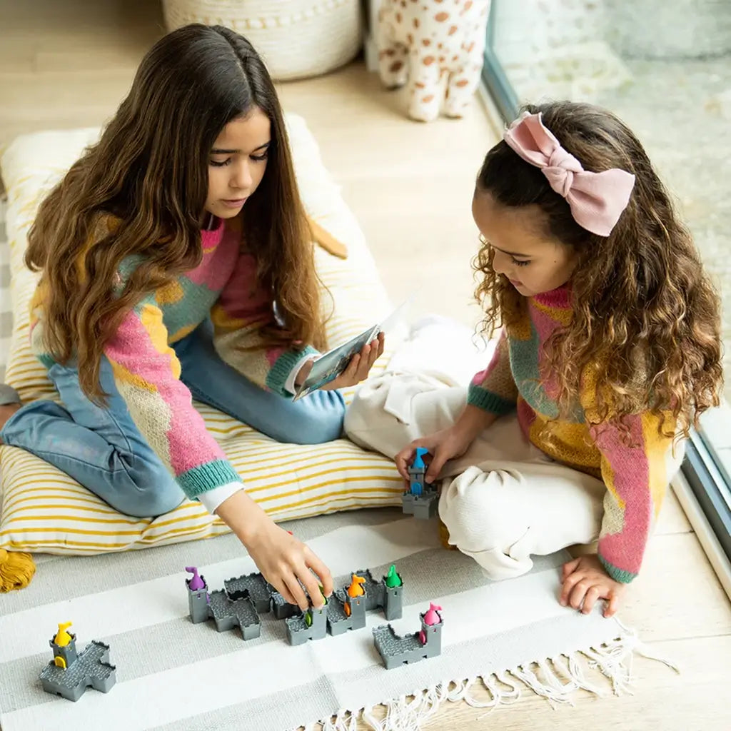 Two young girls play Smart Games Tower Stacks on a striped rug, fitting gray castle pieces with colorful flags together while following instructions from the challenge booklet.