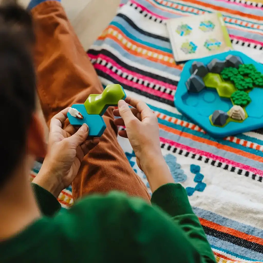 Player assembles Smart Games Treasure Island puzzle while sitting on striped blanket, fitting forest and mountain tiles onto the blue hexagonal board using the challenge guide.
