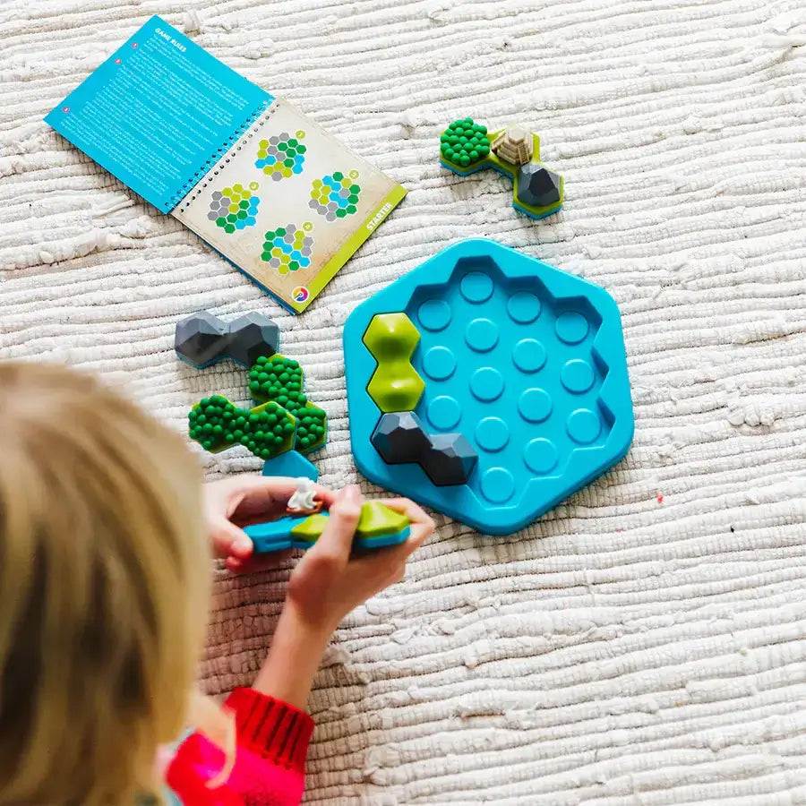 Child sits on textured rug playing Smart Games Treasure Island puzzle, holding landscape tile pieces while following challenges shown in the open instruction booklet.