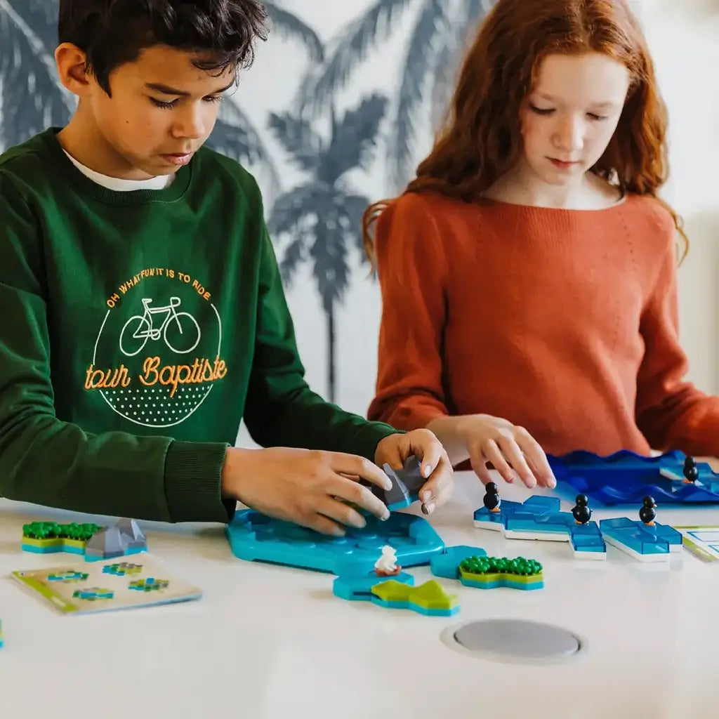 Two children sit at a table playing Smart Games Treasure Island, arranging mountain and forest pieces on the blue puzzle board while referencing the illustrated booklet.