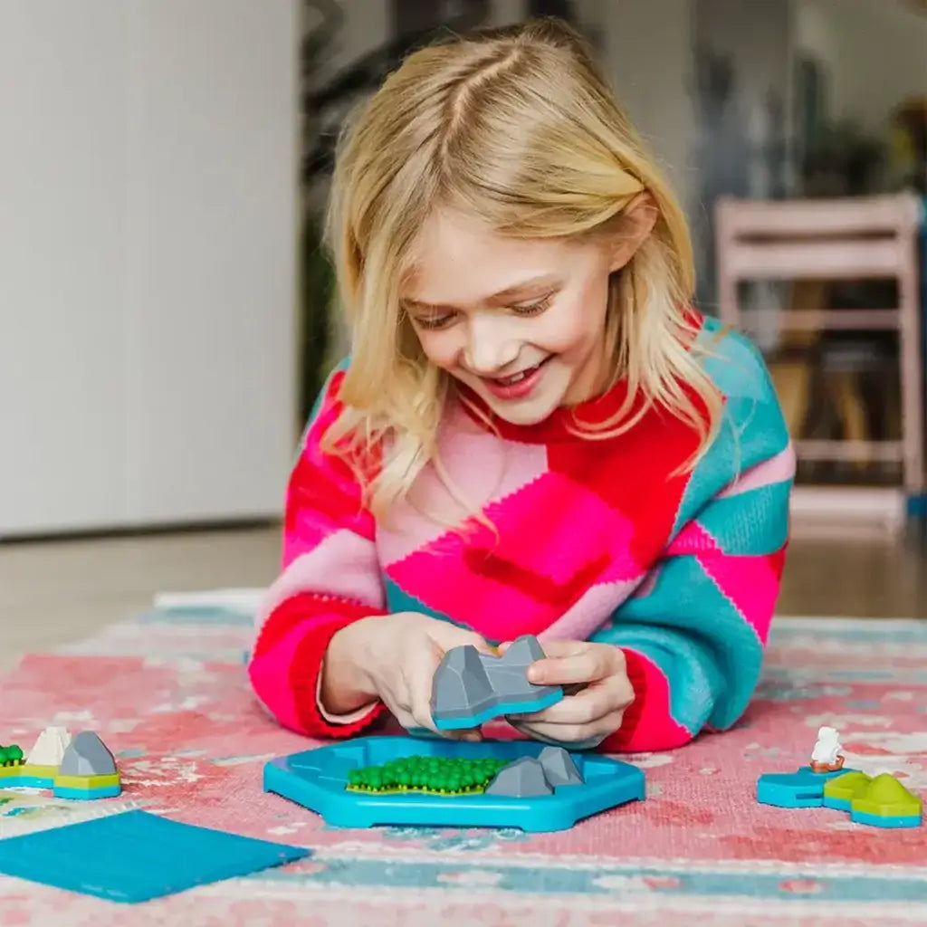 Smiling girl in colorful sweater plays Smart Games Treasure Island, holding a mountain piece above the board while arranging landscape tiles to solve the puzzle.