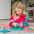 Smiling girl in colorful sweater plays Smart Games Treasure Island, holding a mountain piece above the board while arranging landscape tiles to solve the puzzle.