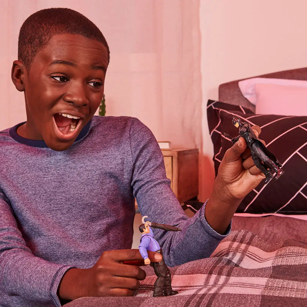 Young boy in a blue long-sleeve shirt smiles widely while playing with DC Spin Master Batman and villain action figures on a neatly made bed with plaid blanket.