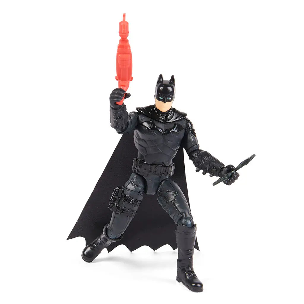 Batman action figure in black armor holds red grappling gun in one hand and black bat-shaped batarang in other, posed in heroic stance with flowing cape.