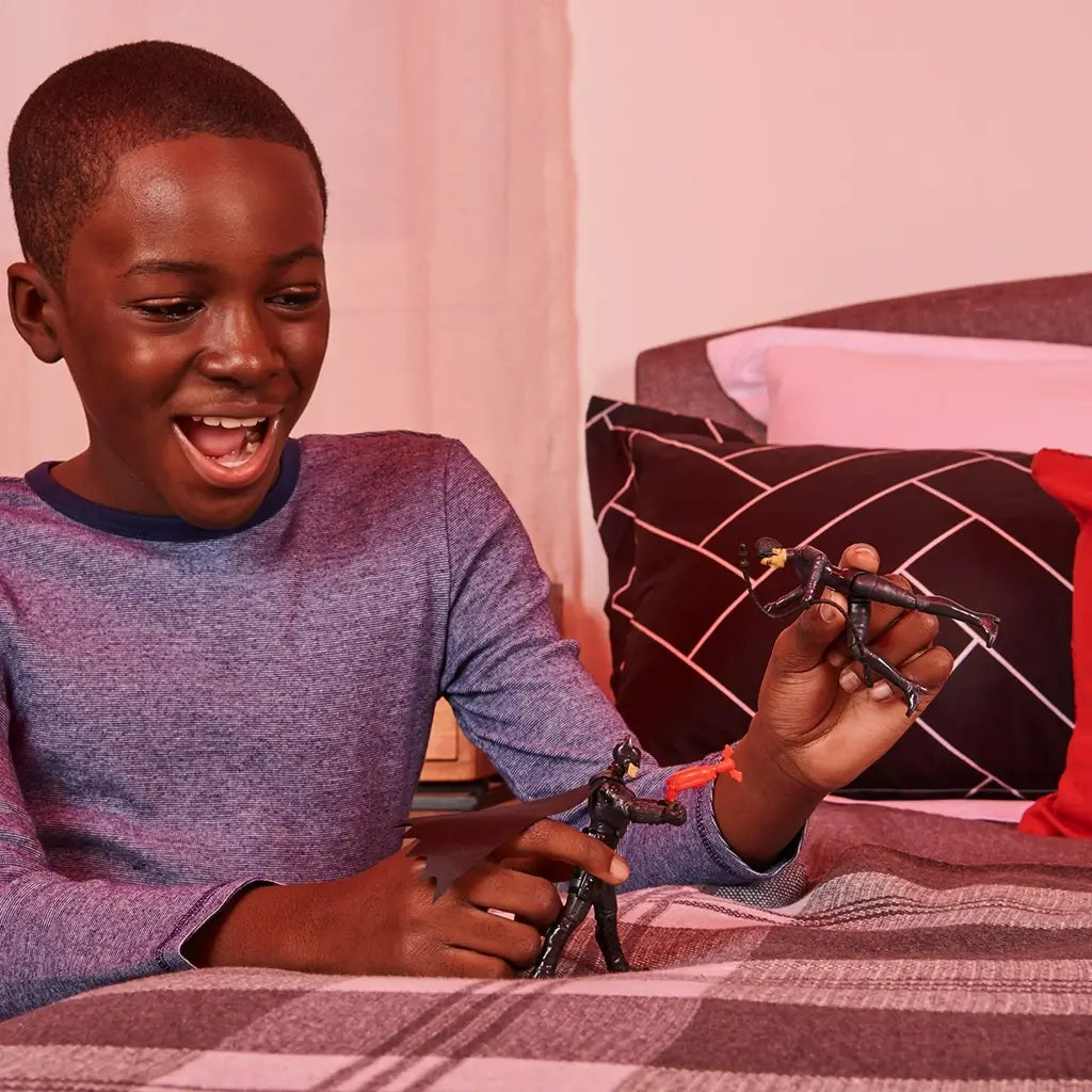 Smiling boy in blue shirt plays on bed with Batman and Selina Kyle action figures, creating a fun battle scene with detailed accessories in a cozy bedroom setting.