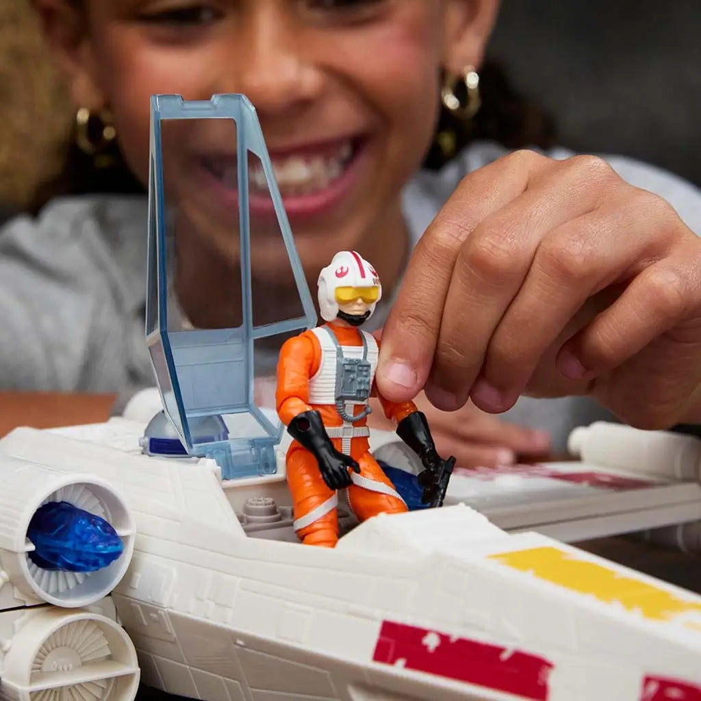 Star Wars Force Strike X-Wing close-up showing cockpit with hinged blue canopy and articulated Luke Skywalker pilot figure in orange flight suit being placed inside by a child’s hand.