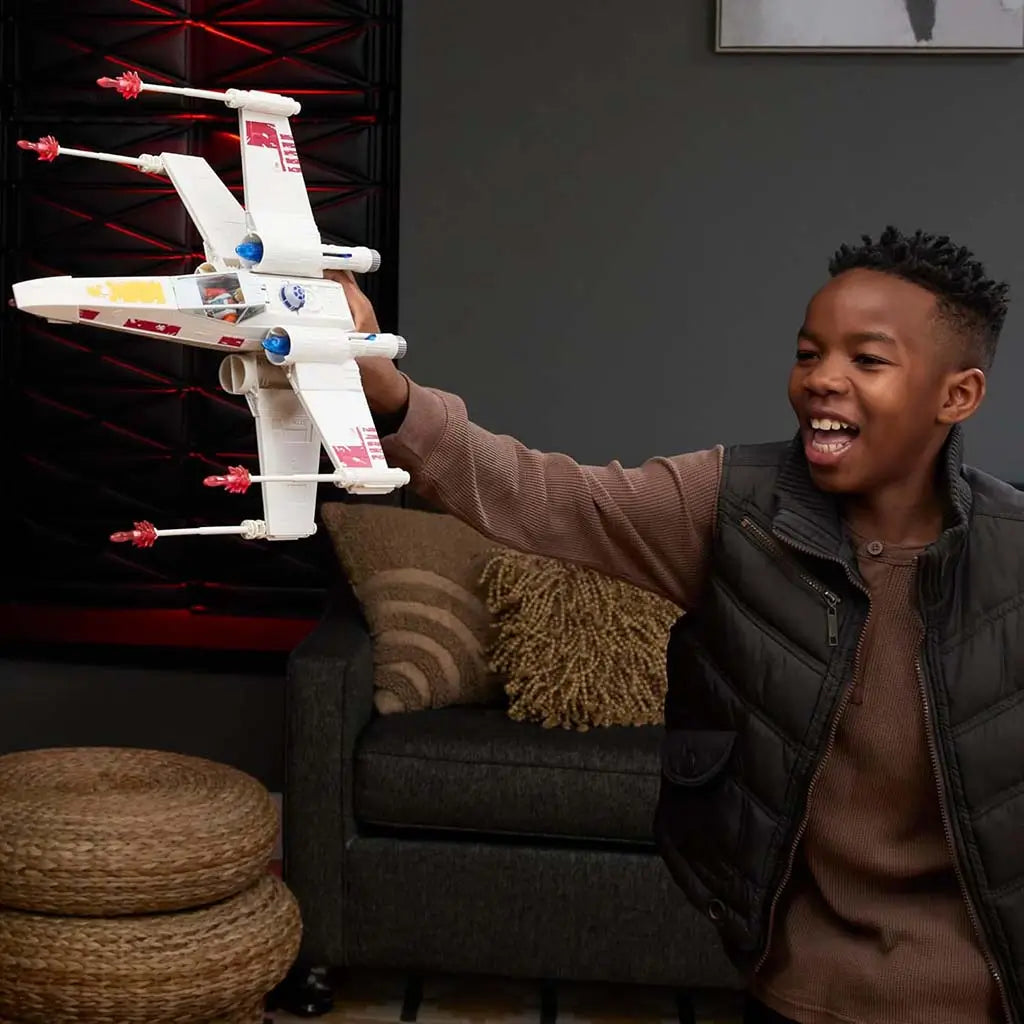 Child playing with Star Wars Force Strike X-Wing toy, holding the white plastic starfighter with red accents and firing-projectile tips while smiling in a modern living-room setting.