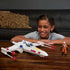 Child smiling at Star Wars Force Strike X-Wing toy resting on wooden table, with white starfighter, red projectile blasts and Luke Skywalker pilot figure arranged for imaginative play.