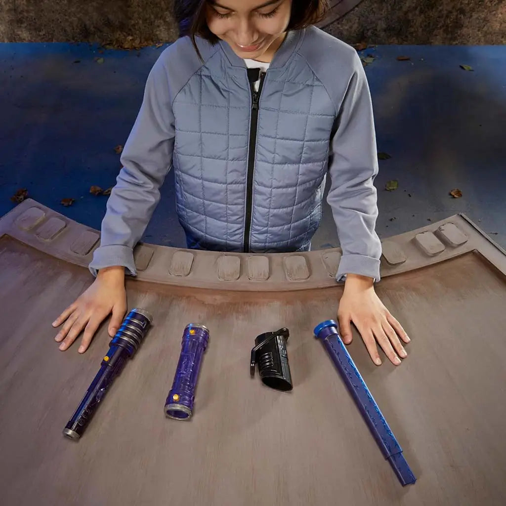 Child arranging Star Wars Lightsaber Forge pieces on a curved wooden surface, including purple hilt parts, black connector, and blue blade, preparing to build a custom lightsaber in a themed play area.