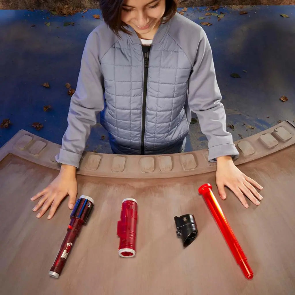 Child arranging Star Wars Lightsaber Forge Darth Vader components on a curved wooden table, including red hilt pieces, black connector, and red blade, preparing the customizable lightsaber for play.