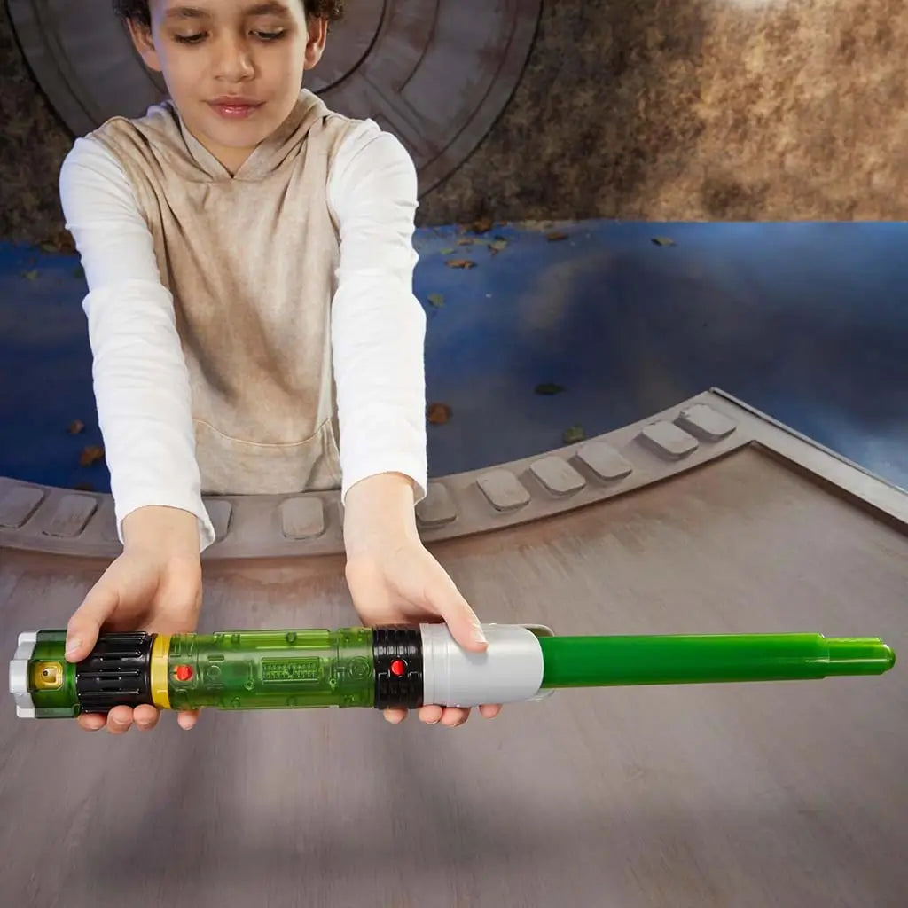 Star Wars Lightsaber Forge Sabine Wren hilt in clear green plastic with black and silver grip, held horizontally by a child assembling the lightsaber on a curved wooden console in a themed setting.