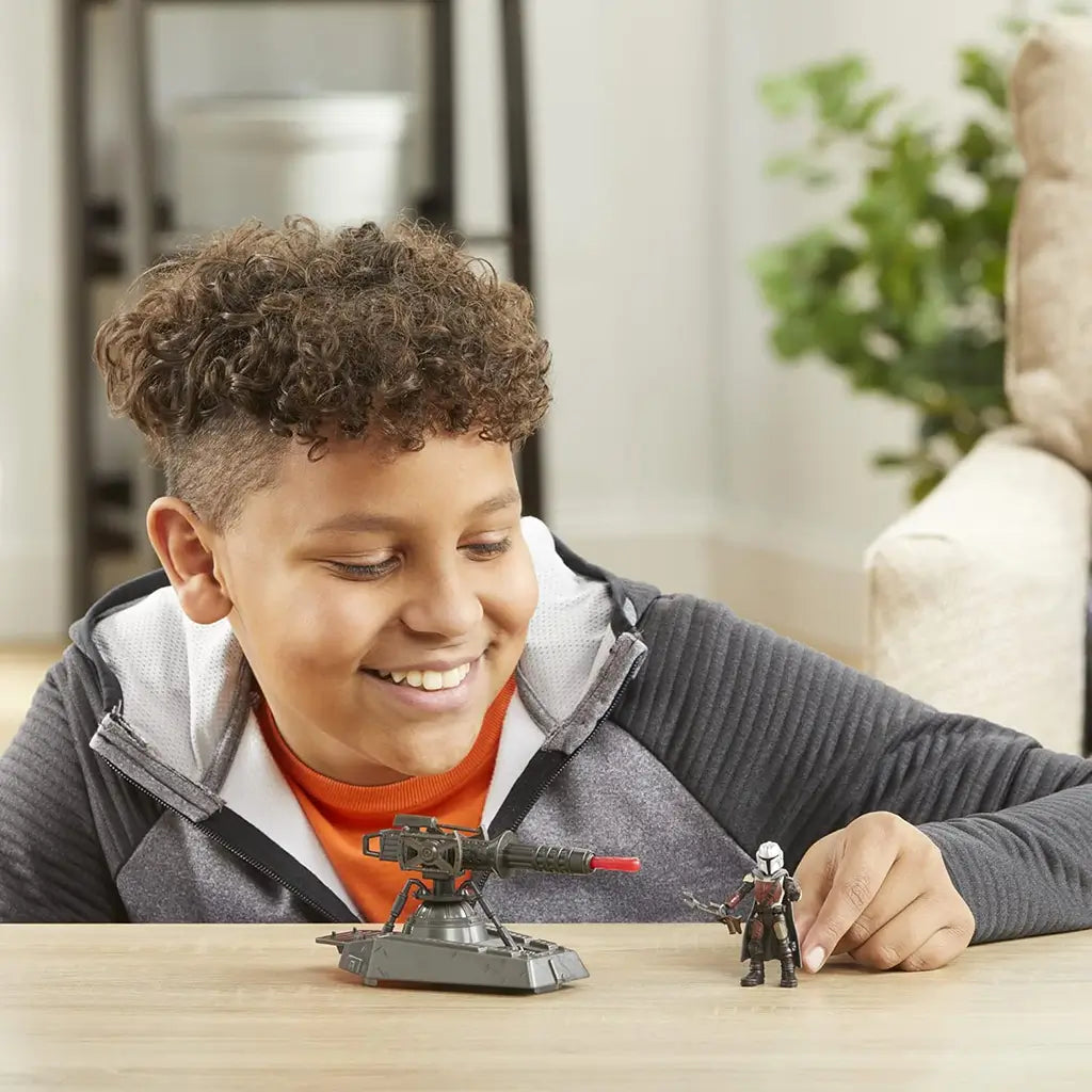 Smiling child in an orange shirt and gray jacket playing with Star Wars Mission Fleet Mandalorian figure and hover E-Web cannon on a wooden table in a cozy, well-lit living room.