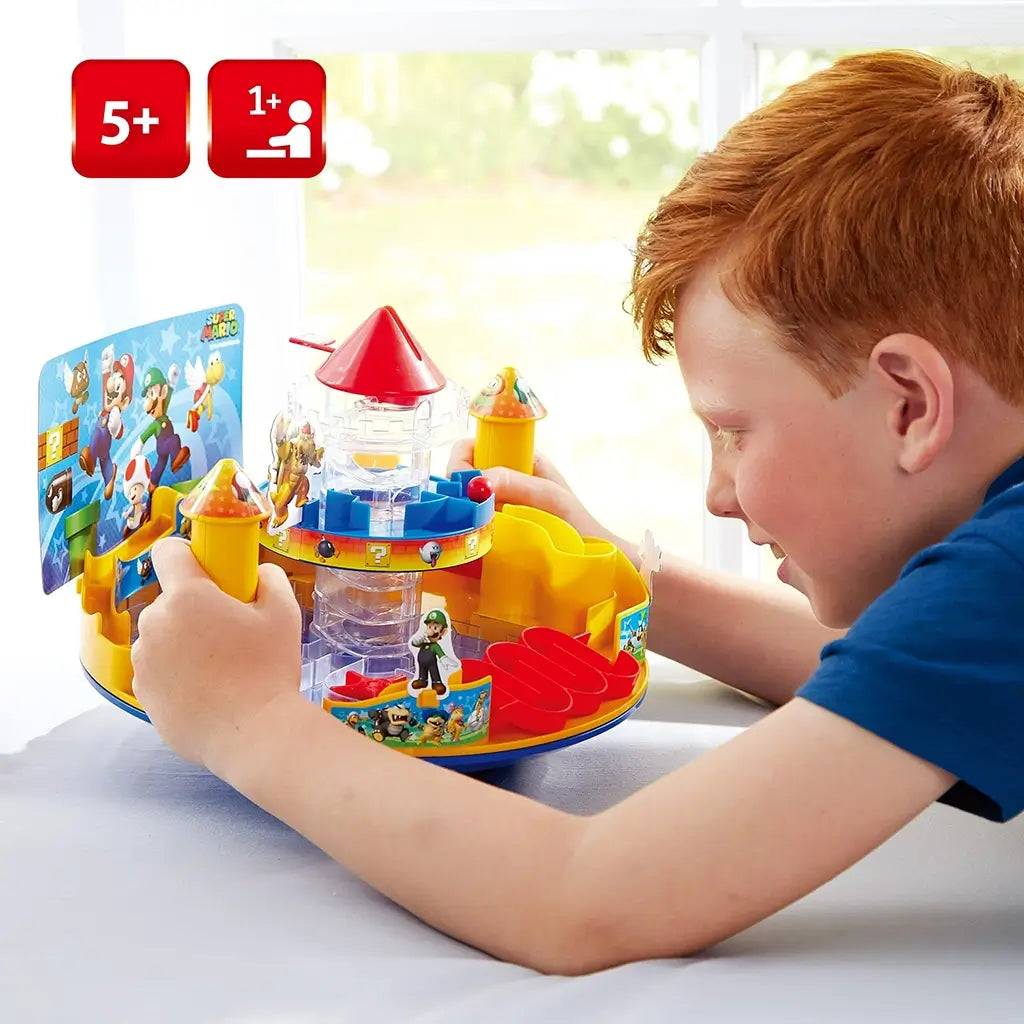 Smiling boy plays Super Mario Castle Land at a table, turning yellow mushroom-shaped controls to guide a ball through the bright plastic maze and interactive castle tower traps.