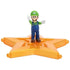 Super Mario Galaxy Movie Luigi figure wears green and blue overalls with white gloves and brown shoes, posed with both arms raised on a glossy orange-gold star platform.