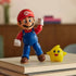 Super Mario Galaxy Movie Mario figure features red and blue articulated plastic, posed with one fist raised beside a smiling yellow Luma accessory atop stacked books indoors.