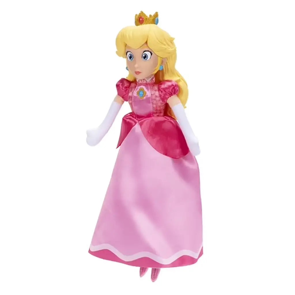 Princess Peach plush toy from The Super Mario Galaxy Movie has a soft pink satin gown, blonde hair, gold jeweled crown, white gloves, and blue earrings on a white background.