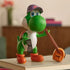 Super Mario Galaxy Movie Yoshi figure has green textured plastic and orange boots, wearing a purple New York City cap while holding sunglasses beside an orange bag on books.