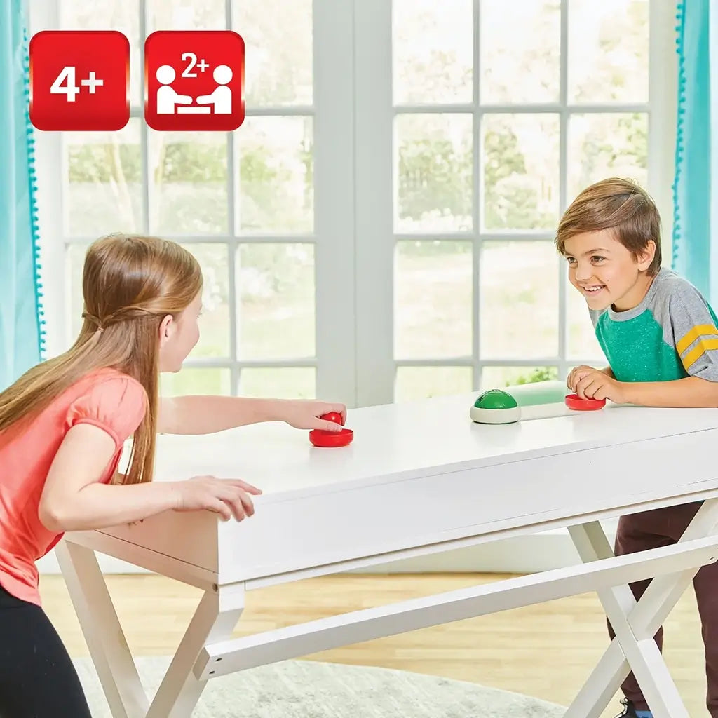 Two children laughing while playing Super Mario Hover Shell Strike at a white table, each holding red strikers while a green shell puck hovers between them indoors.