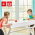 Two children laughing while playing Super Mario Hover Shell Strike at a white table, each holding red strikers while a green shell puck hovers between them indoors.