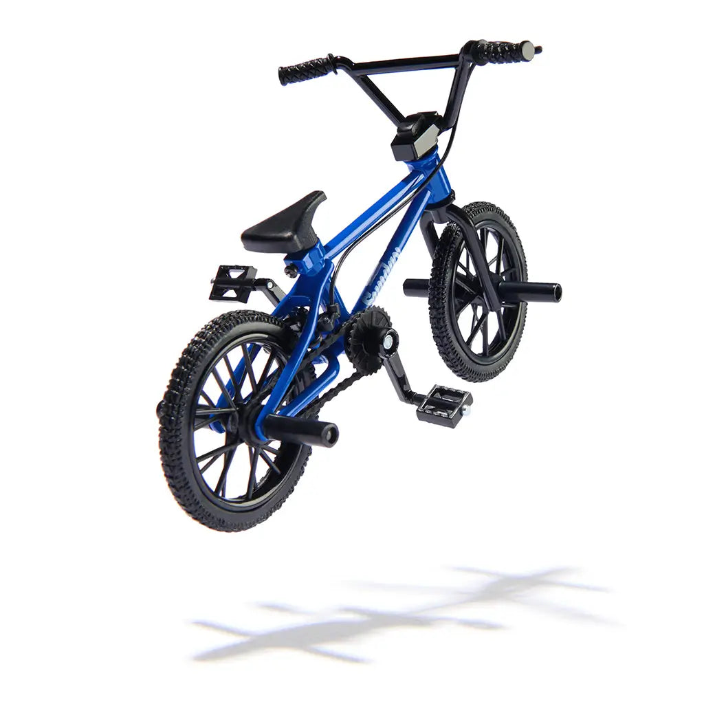 Tech Deck BMX finger bike in blue and black metal and plastic, compact stunt frame with spoked wheels, rear pegs, and pedals, designed for tricks, shown in midair on a minimal white background.