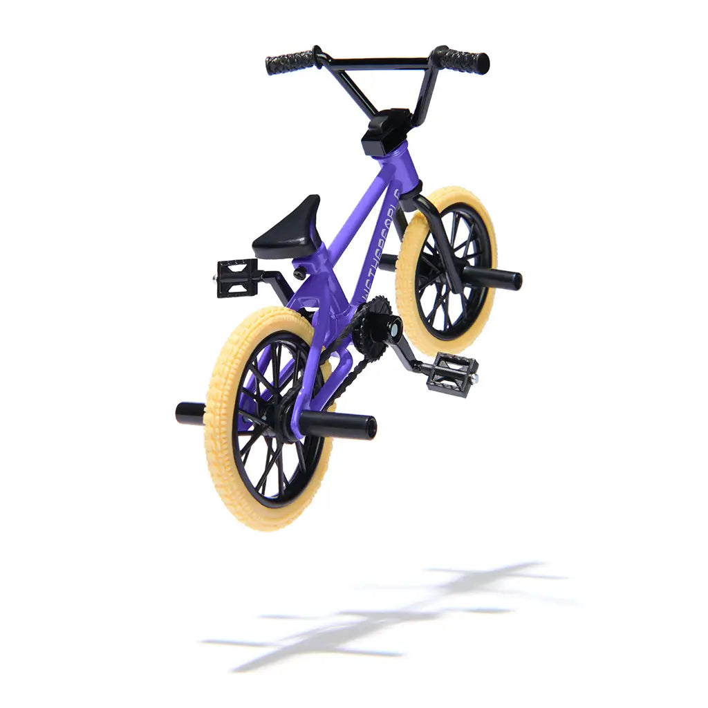 Tech Deck BMX finger bike in purple metal frame with black components and tan tires, compact freestyle stunt bike form with rear pegs and pedals, displayed midair on a clean white background.