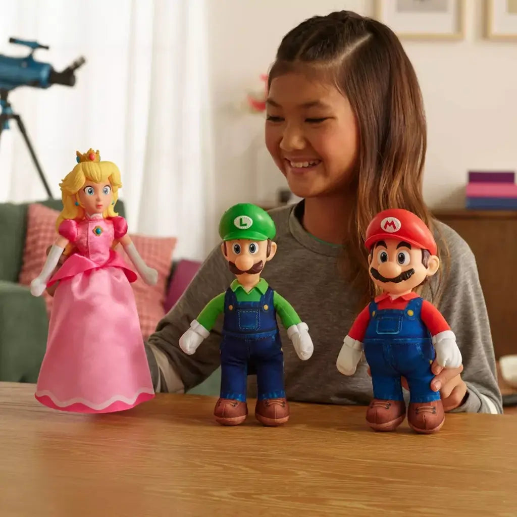 Super Mario plush toys feature colorful fabric characters including Luigi in green and Mario in red with Princess Peach in pink dress, soft textile texture, displayed with child playing at table in cozy indoor setting.