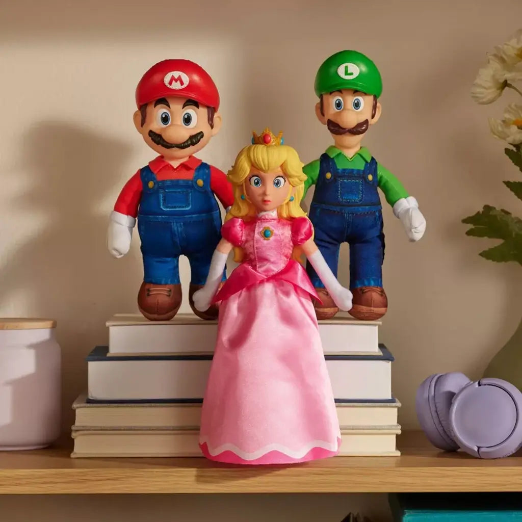 Super Mario plush toy set shows Princess Peach in pink gown with Mario and Luigi, soft textile material and smooth stitched details, arranged on stacked books with cozy home decor background.