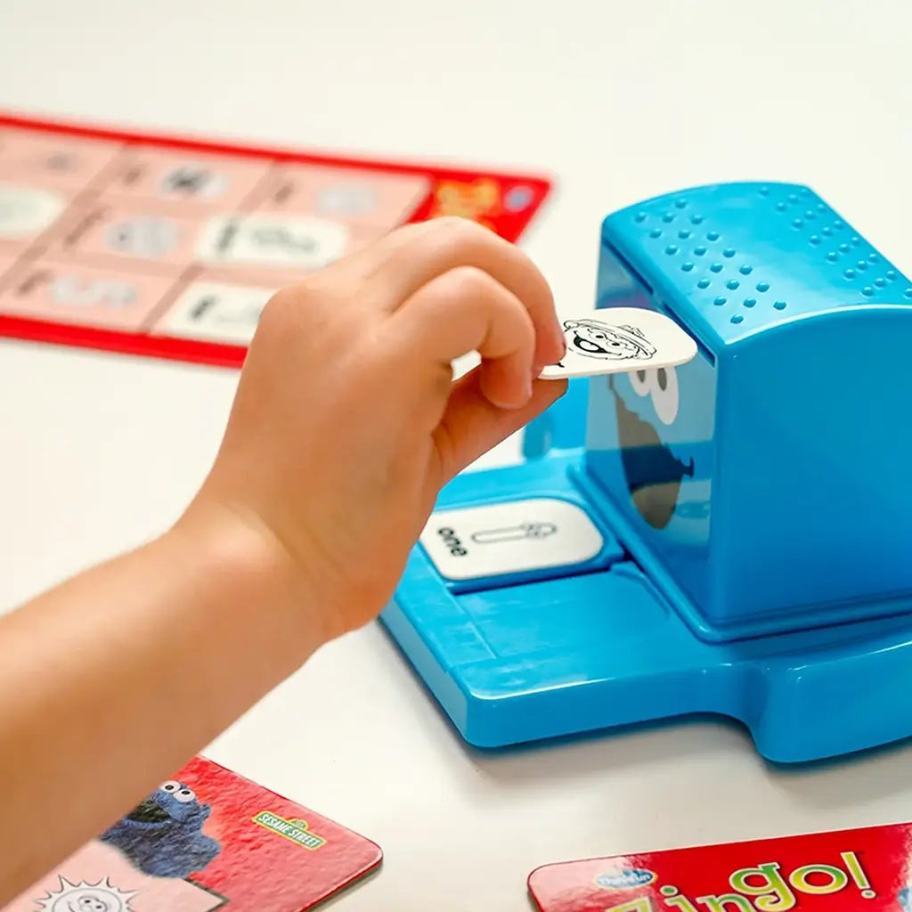 Close-up of ThinkFun Sesame Street Zingo gameplay showing a child placing a black-and-white picture tile into the Cookie Monster Zinger dispenser, with bingo cards spread nearby.
