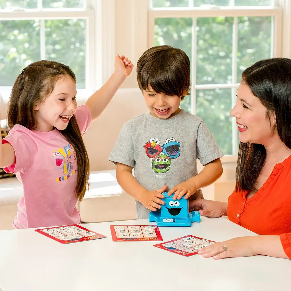 Two children and an adult playing ThinkFun Sesame Street Zingo at a table, with kids smiling and using the Cookie Monster Zinger while matching tiles to colorful bingo cards in play.