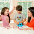 Two children and an adult playing ThinkFun Sesame Street Zingo at a table, with kids smiling and using the Cookie Monster Zinger while matching tiles to colorful bingo cards in play.