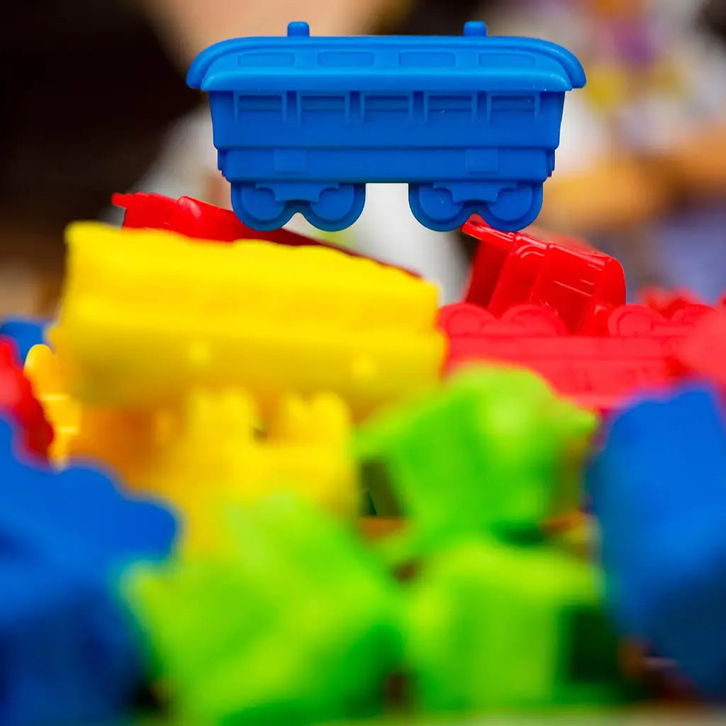 Ticket to Ride First Journey train pieces feature bright blue, red, yellow, and green molded plastic rail cars, with one blue car sharply focused against a blurred game setting.