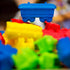 Ticket to Ride First Journey train pieces feature bright blue, red, yellow, and green molded plastic rail cars, with one blue car sharply focused against a blurred game setting.