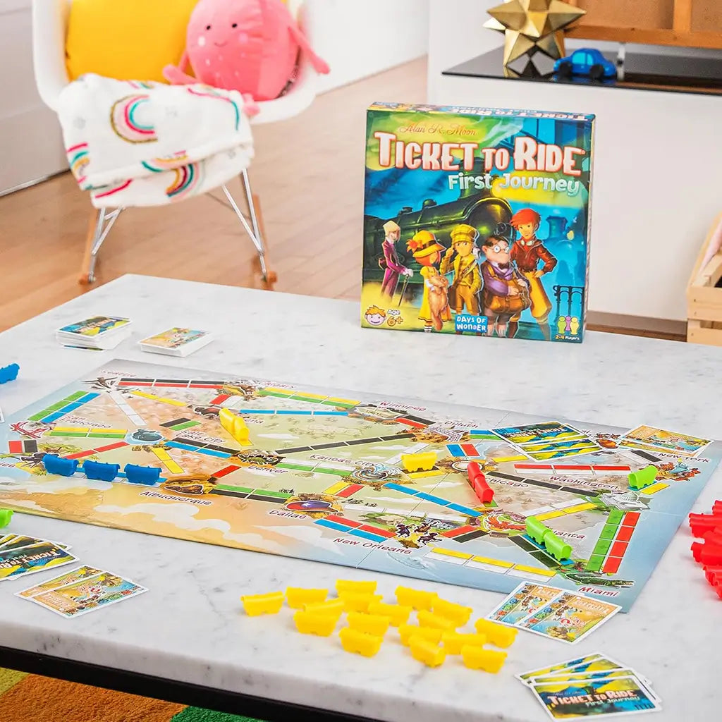 Ticket to Ride First Journey board game displays a colorful North America route map with yellow, blue, green, and red trains, cards, and the game box on a white table.