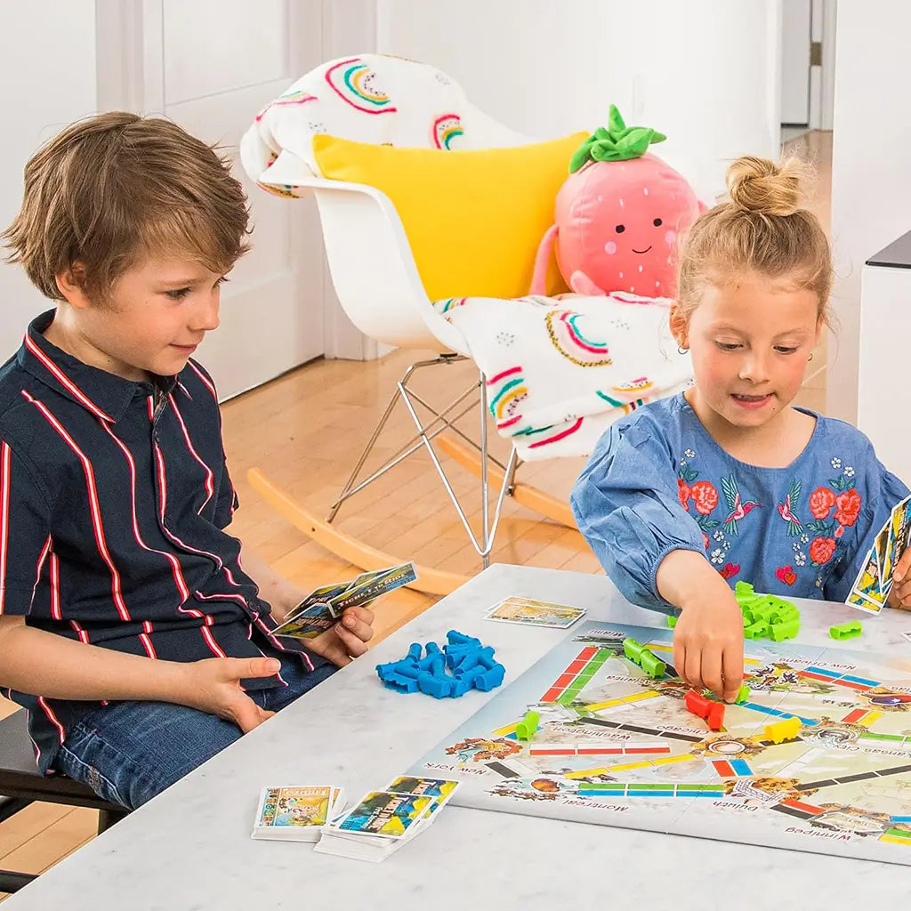 Ticket to Ride First Journey kids board game shows a boy and girl holding cards and placing colorful plastic trains on the North America route map at a white table.