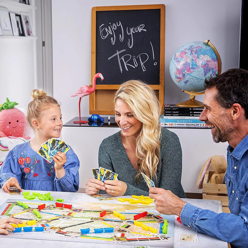 Ticket to Ride First Journey family board game shows a girl and two adults holding cards around a colorful train route board in a bright playroom with travel-themed decor.