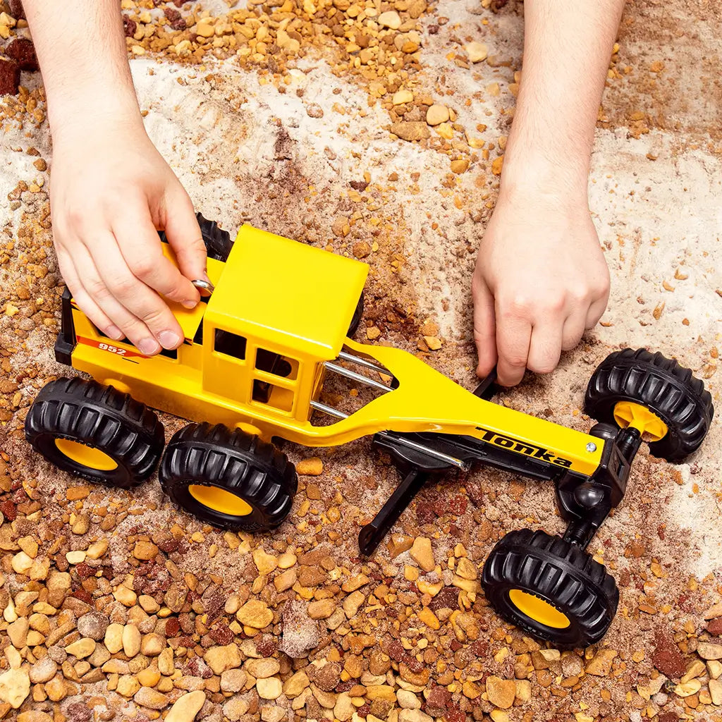 Hands playing with yellow Tonka grader toy on sandy, rocky terrain, showcasing the toy’s rugged tires and durable build for outdoor play.