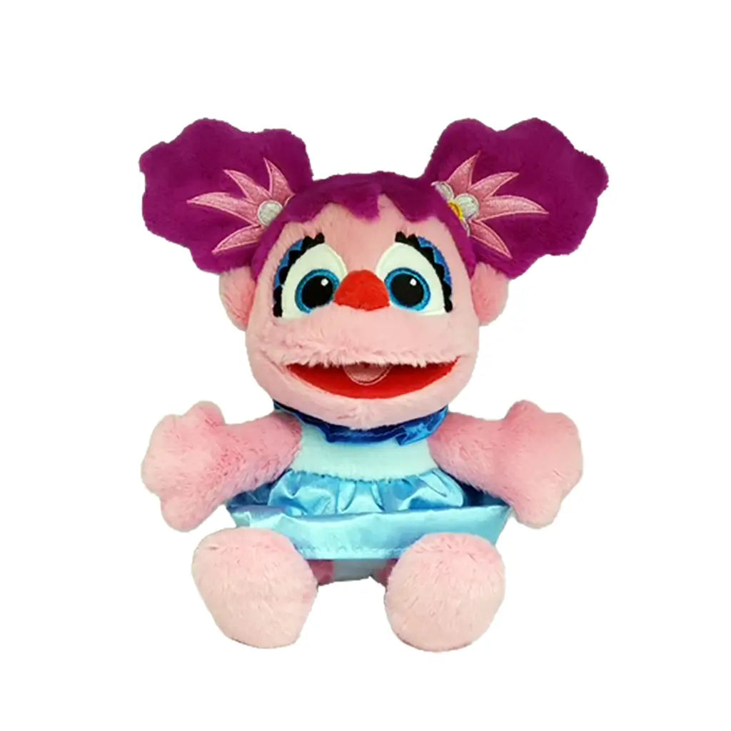 Sesame Street Abby Cadabby plush toy in pink fuzzy fabric with rounded character shape, featuring bright blue eyes, purple pigtails, and a light blue dress, designed as a soft collectible doll on a white background.