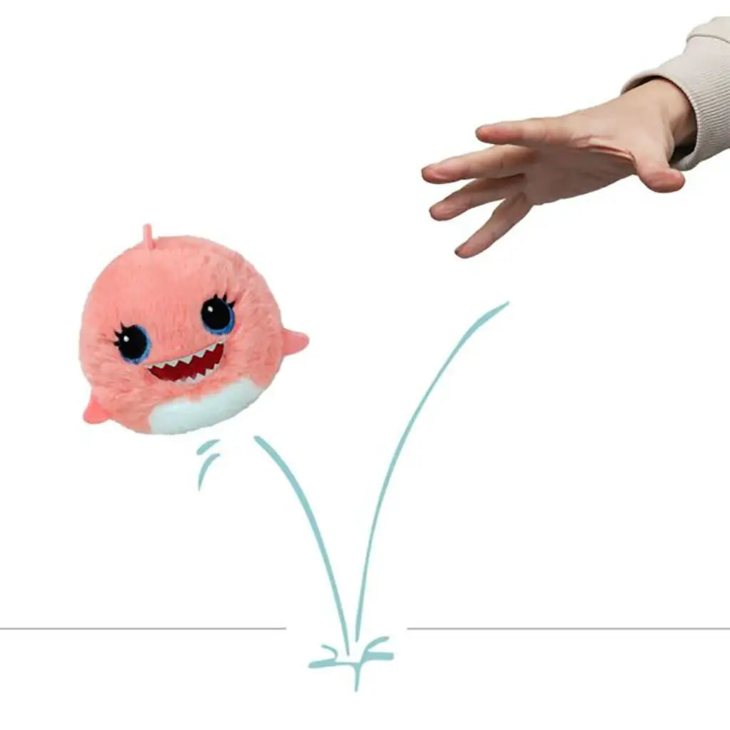 Plush toy Reef the Pink Shark by Ty Beanie Bouncers, shown bouncing with a hand releasing it, round fuzzy pink body, white belly, embroidered blue eyes, smiling teeth design.