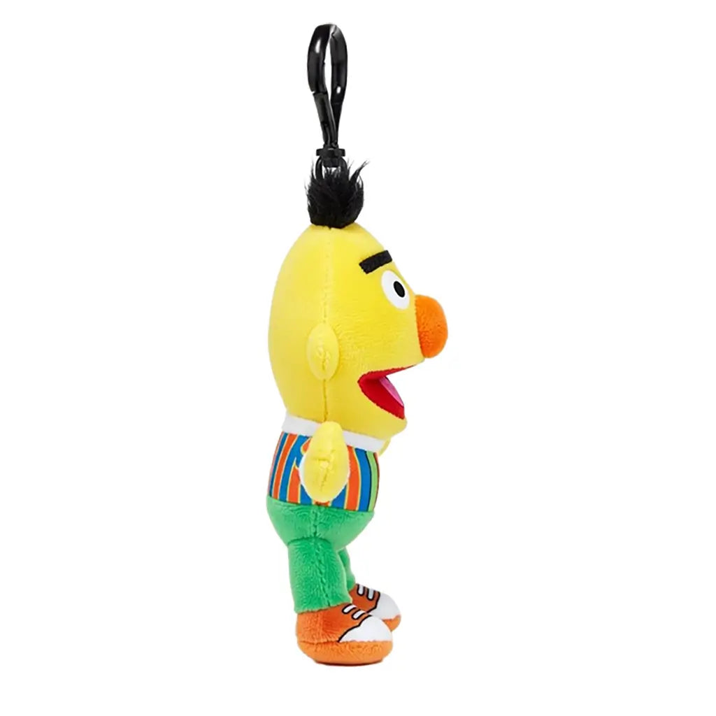 Sesame Street Bert plush clip in yellow soft fabric, with black hair, orange nose, striped shirt, green pants and orange shoes, shown in side view with a black hook on white.
