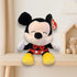 A smiling TY plush Mickey Mouse with closed eyes sits on a light wooden shelf in a cozy nursery setting, with a teddy bear in a small house-shaped shelf and a cloud wall decoration in the background.