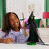 Wicked Elphaba fashion doll lifestyle scene shows a smiling child playing indoors, with the green-skinned Elphaba doll posed on a stand holding a broom, demonstrating scale, poseability, and imaginative play in a colorful bedroom setting.