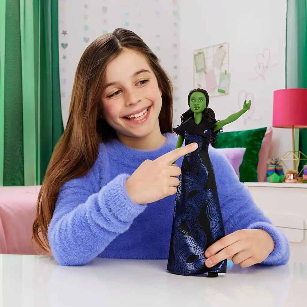 Wicked Elphaba singing doll shown in a lifestyle play scene with a smiling child, green-skinned doll in patterned gown held upright, illustrating interactive singing play in a bright bedroom setting.