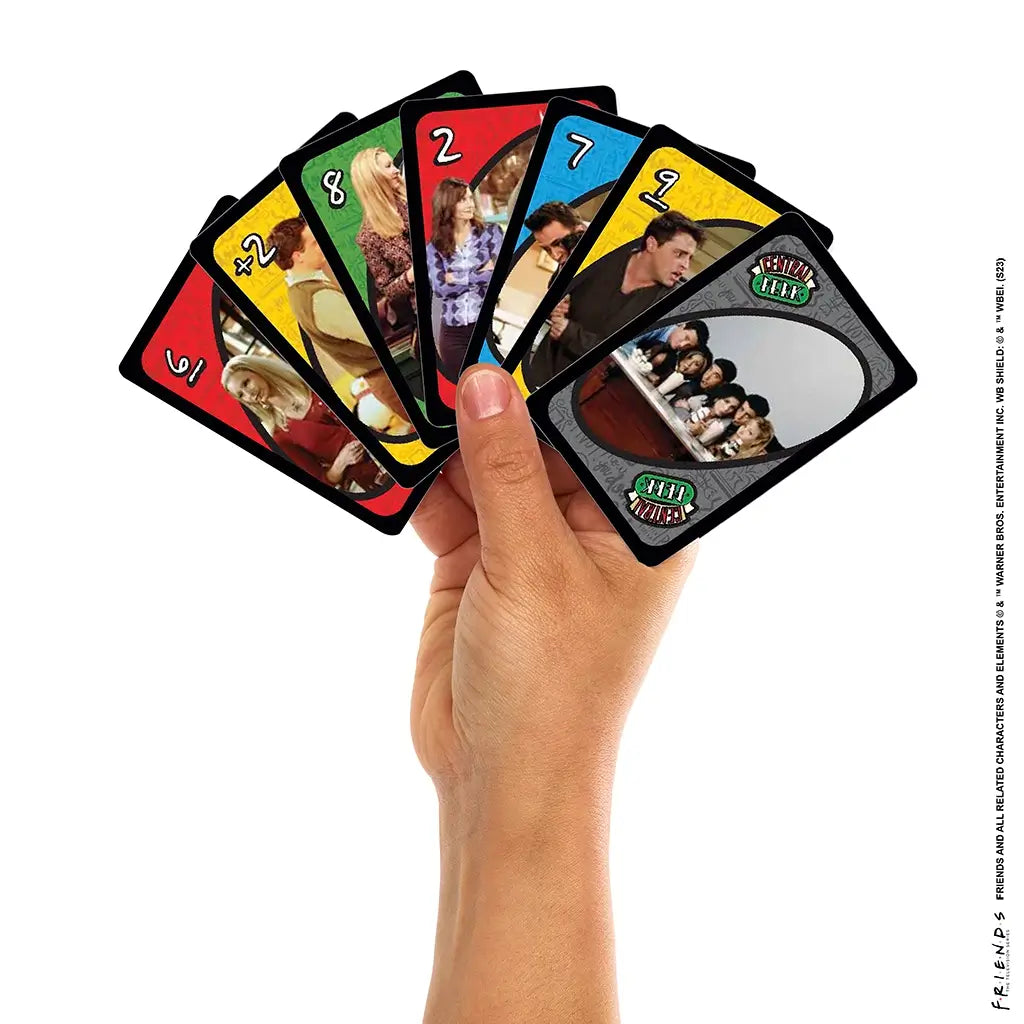 A hand holds a fan of UNO Friends cards featuring colorful numbers and TV show moments with Chandler, Monica, Phoebe, and the group sitting at Central Perk.