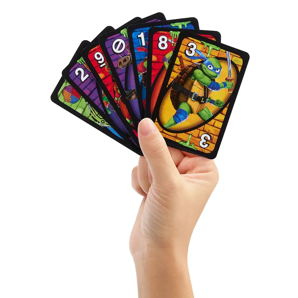 Hand holding a fan of UNO Teenage Mutant Ninja Turtles Mutant Mayhem cards, displaying turtle characters, colorful numbers, and special action cards against a white background.