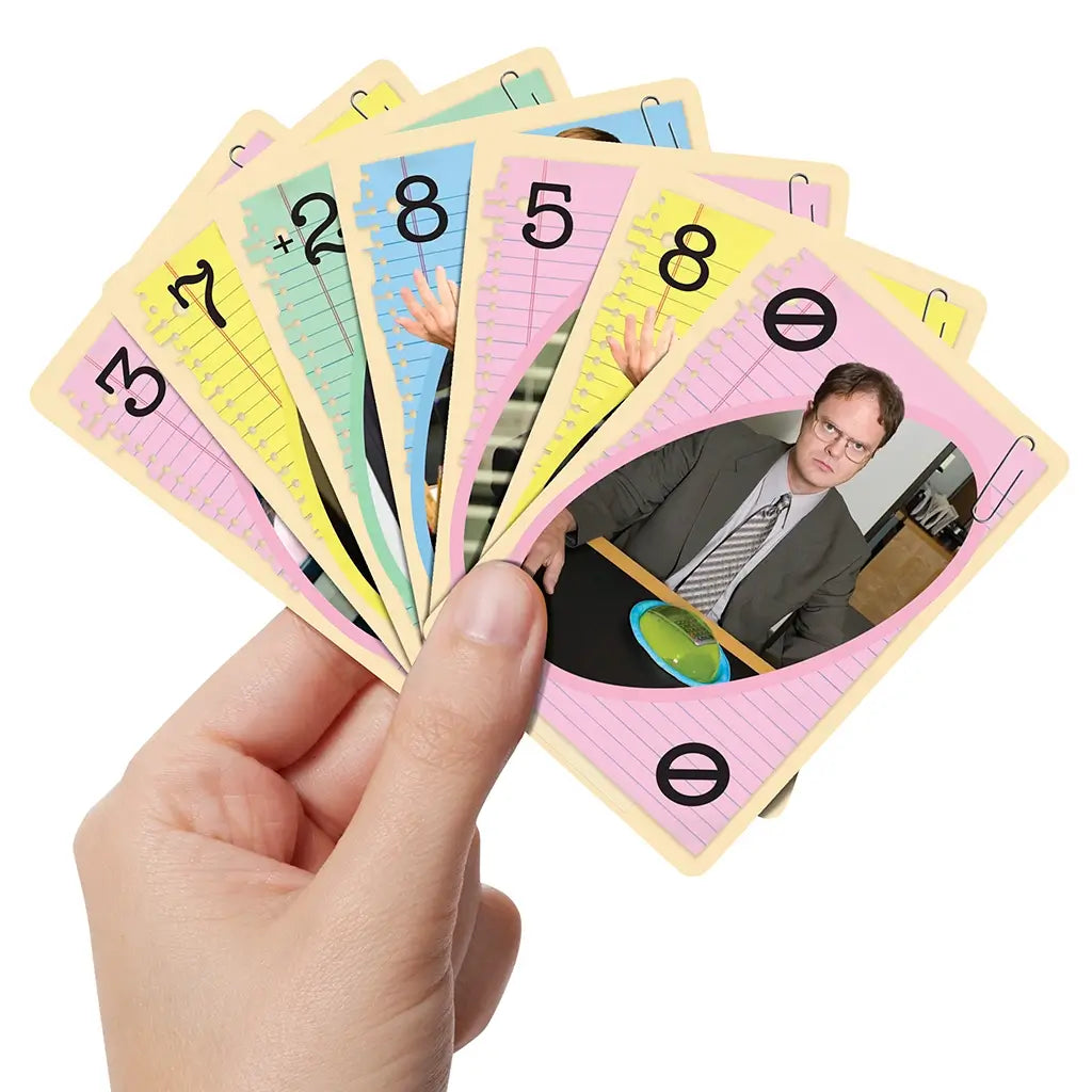 Hand holding a fan of UNO The Office cards showing pastel backgrounds with character photos including Dwight and action cards, styled with paperclip and notebook page themes.